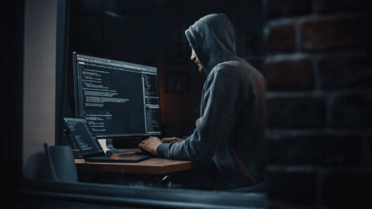 Silhouette encapuchonnée devant un écran de code dans une pièce sombre